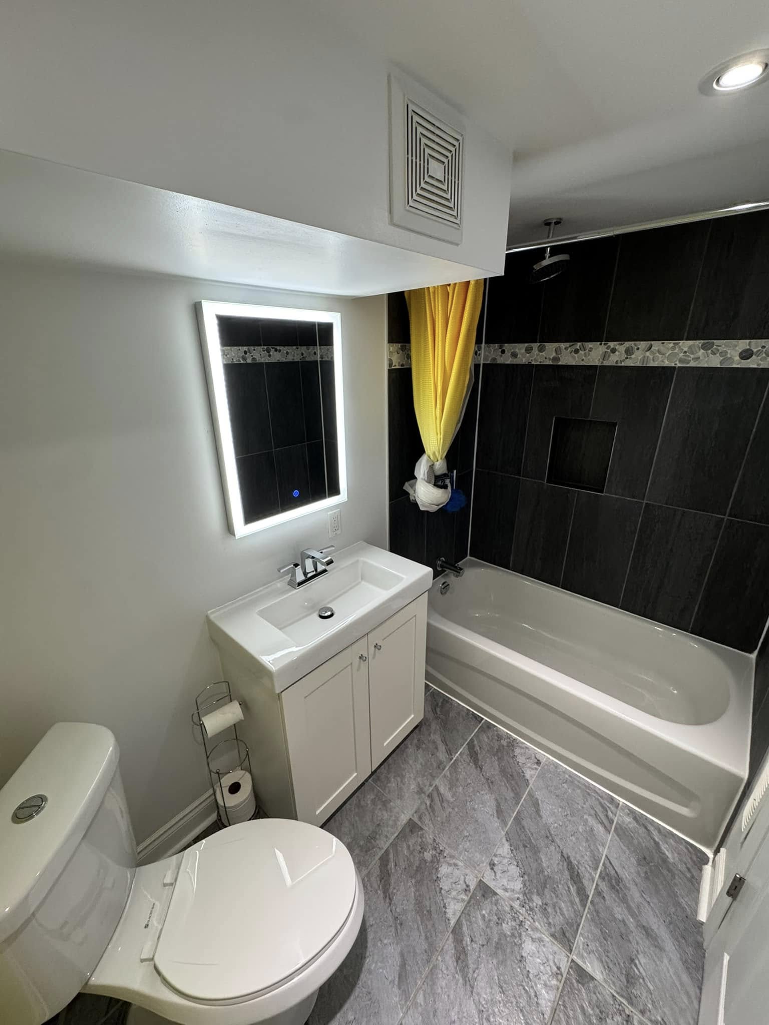 Bathroom with black tiled tub surround, backlit mirror and white vanity.