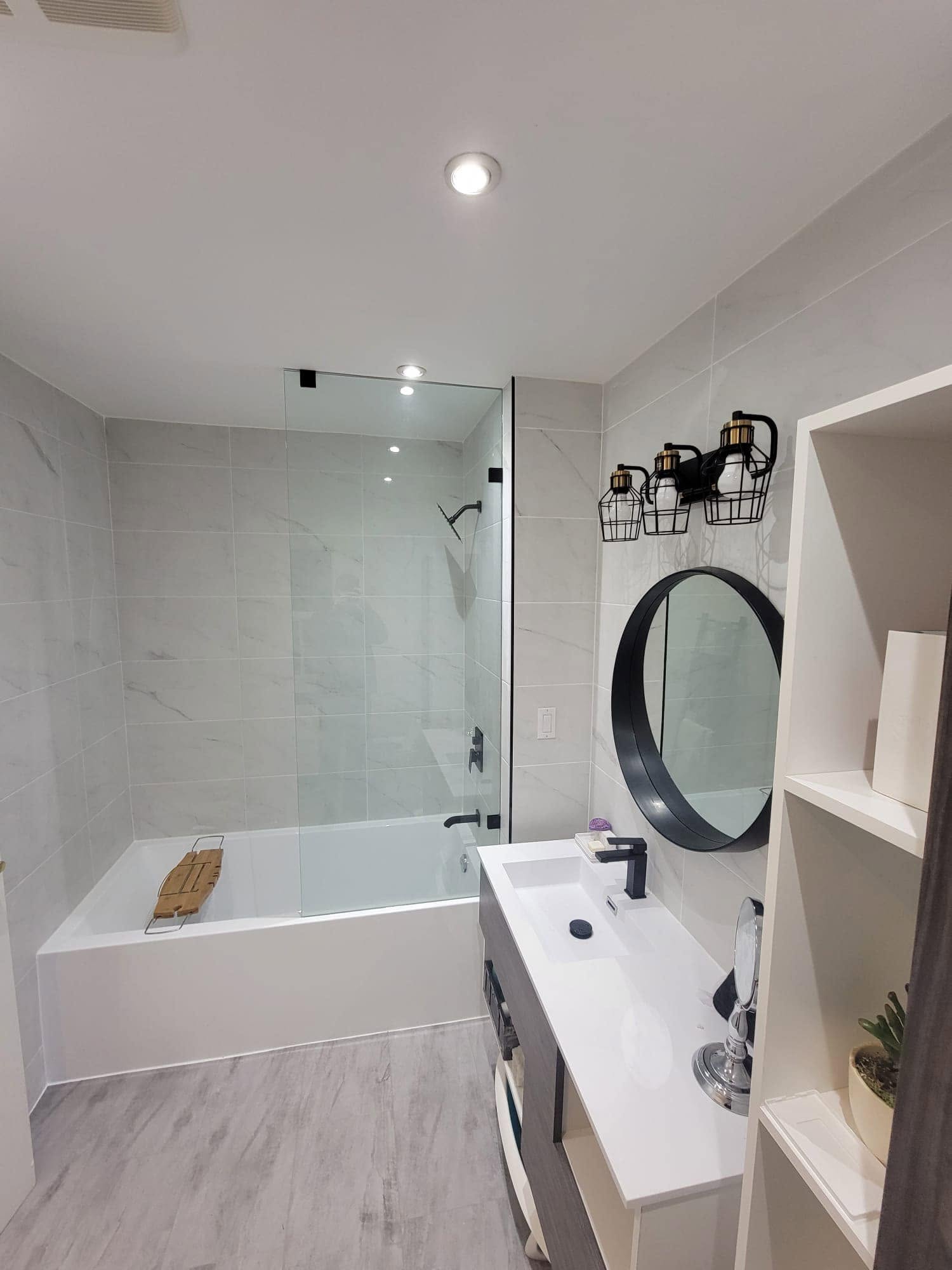 Bathroom with a tub behind a glass screen, round black mirror and white vanity.