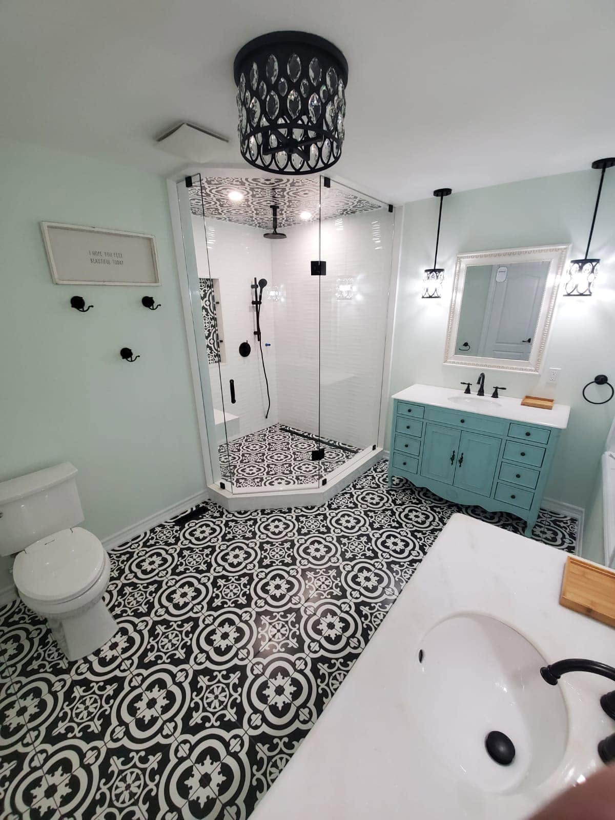 Bathroom with patterned black and white floor tile, glass corner shower and teal vanity.